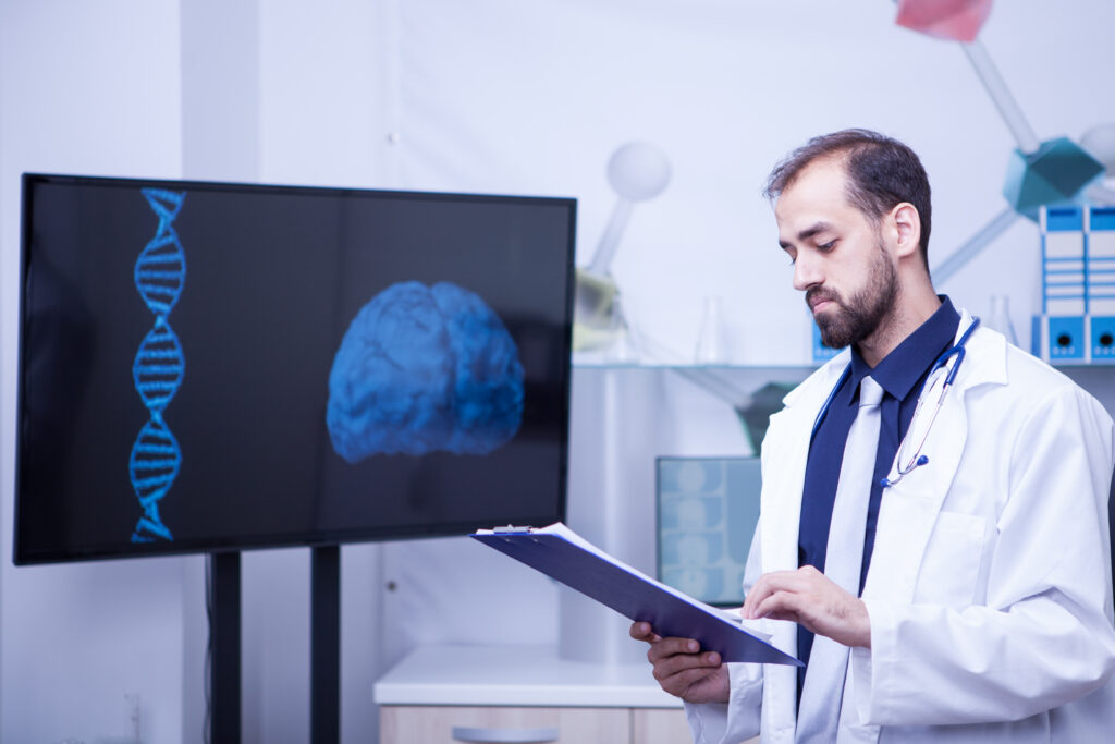 Confident young doctor with a clipboard in his hands standing by a monitor with a brain graph