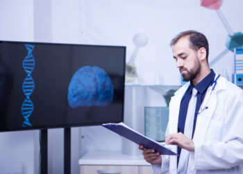 Confident young doctor with a clipboard in his hands standing by a monitor with a brain graph