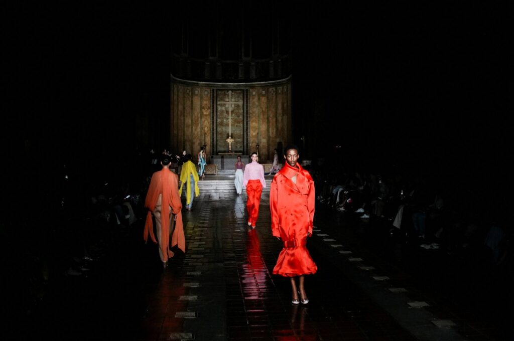 models in red, orange, yellow and pink walking a dark church nave during NYFW