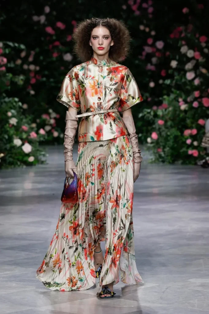 Model in a Luisa print mandarin-collar blouse belted over a pleated floral skirt, long taupe leather gloves, and a small glass bag