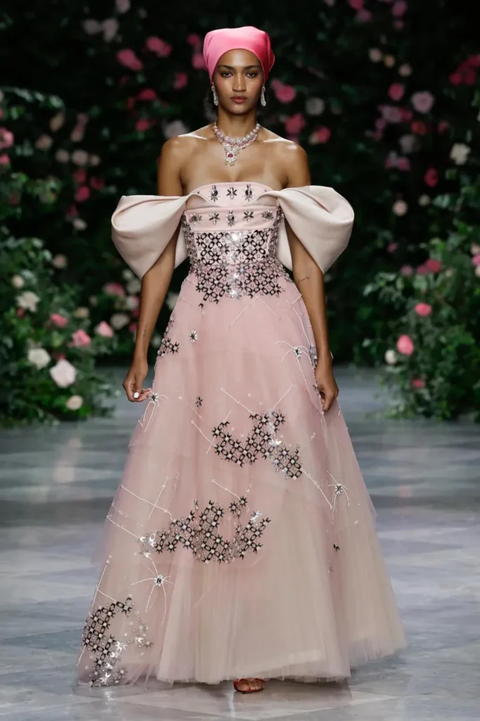 Model in a blush tulle ball gown with sculpted satin off-shoulder sleeves, black and silver geometric embroidery, and a pink satin headscarf