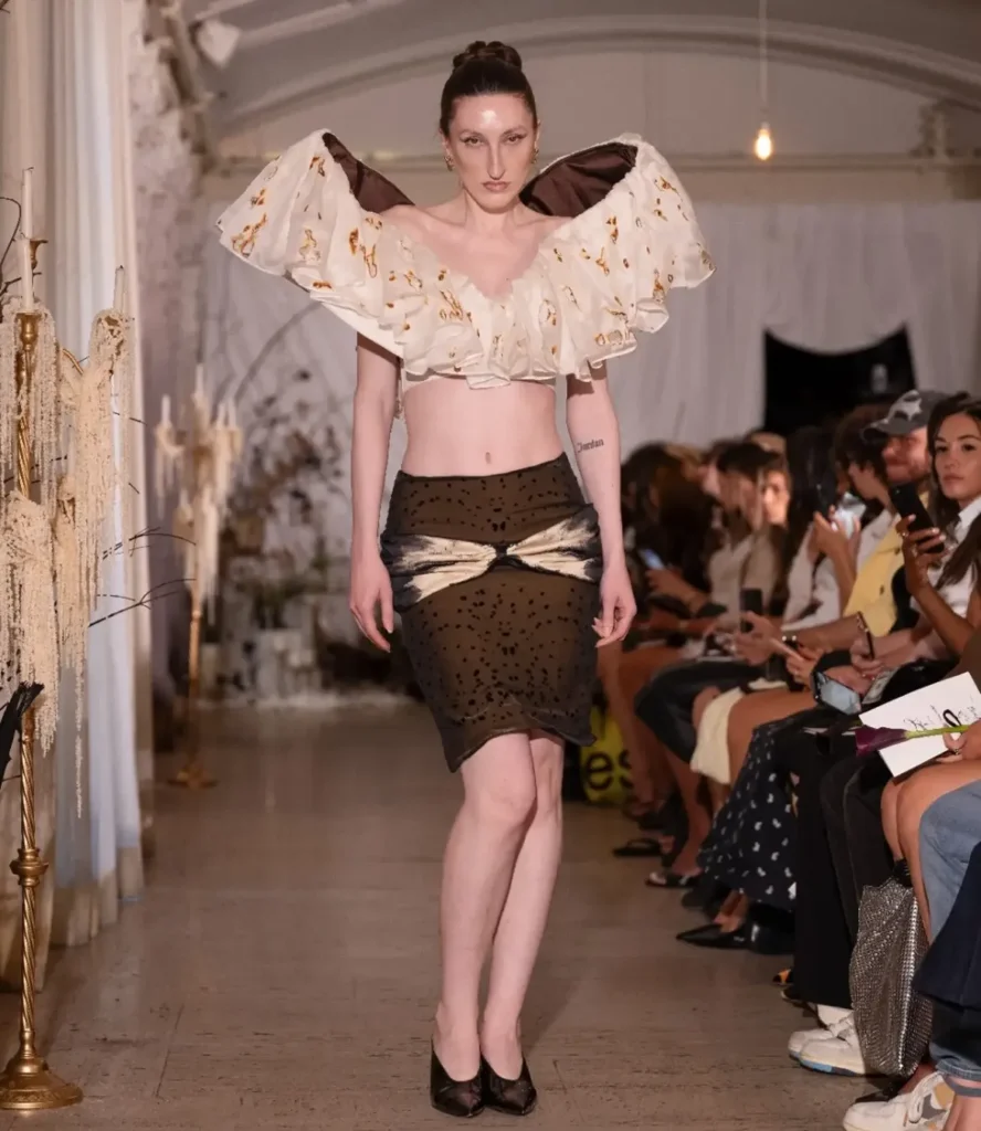 Model in a cropped top with oversized ruffled wing shoulders in ivory with amber marks, over a brown spotted pencil skirt