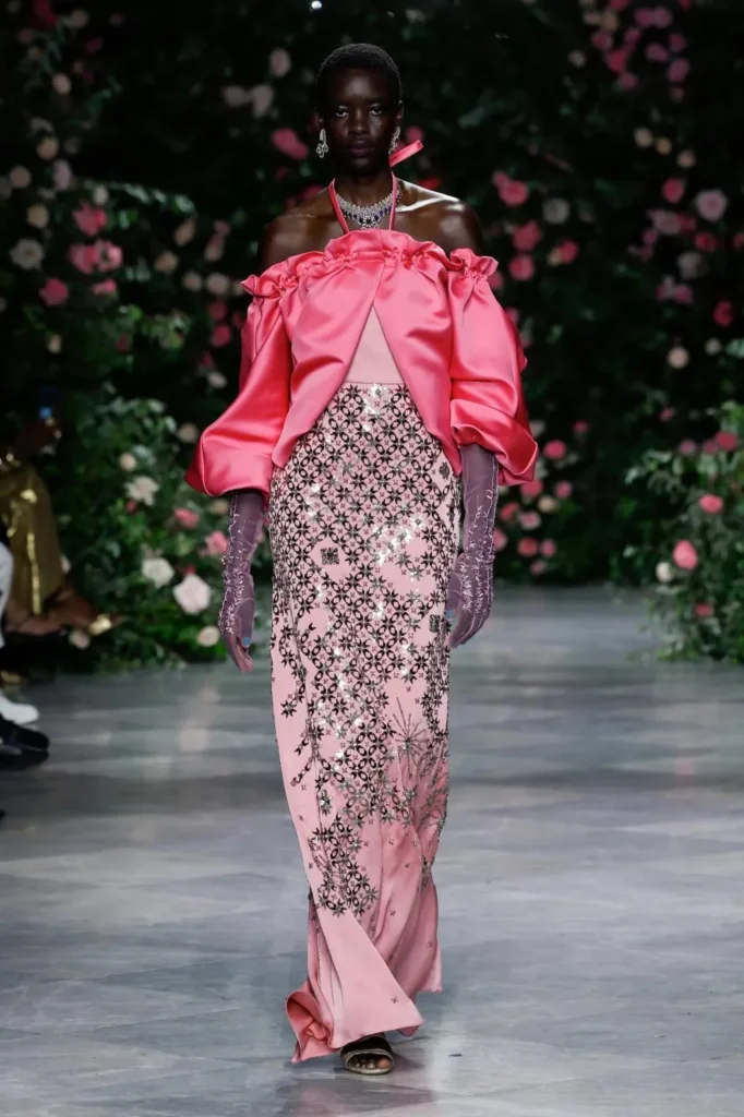 Model in a hot-pink satin ruffled off-shoulder jacket over a pale pink column gown with black and silver lattice embroidery and lilac embroidered gloves