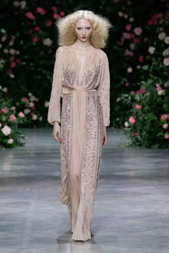 Model in a mustard-gold chiffon gown with billowing sleeves, white vine embroidery in vertical lines, pleated skirt, and a feathered corsage at the shoulder