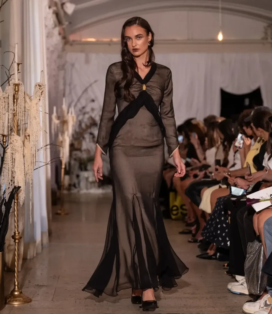 Model in a sheer bronze long-sleeved chiffon gown with a black chiffon drape crossed at the waist and a gold brooch