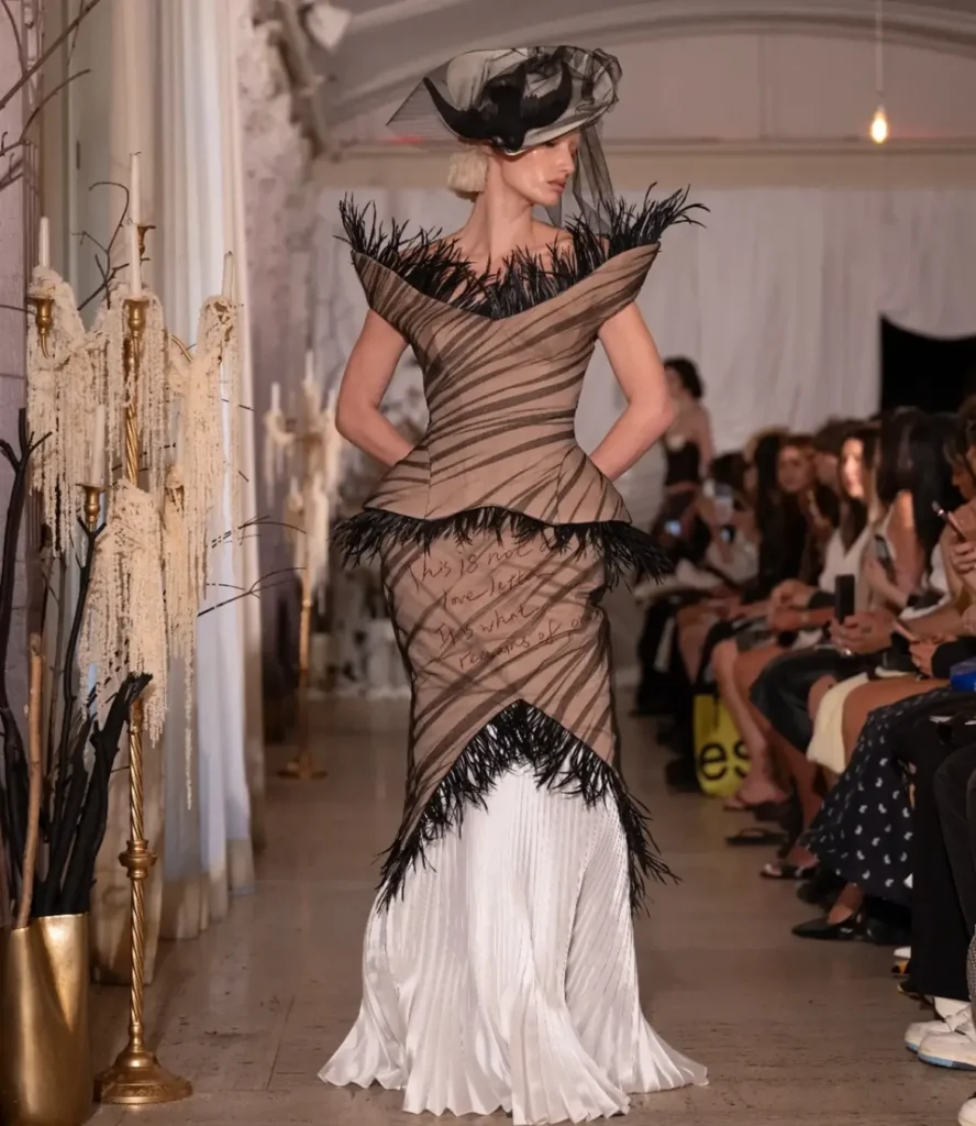 Model in a taupe pleated tulle gown with black feather trim, handwritten text across the skirt, and a black veiled feather hat.