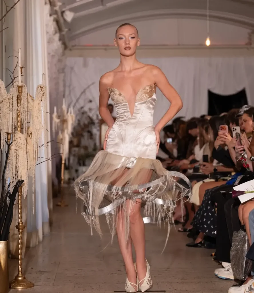 Model in an ivory satin corset dress with a sheer fringed hoop skirt exposing the ribbon frame