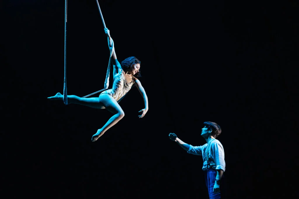 the full company under the blue big-top canopy, aerialists suspended overhead, hoops and rope work across the stage