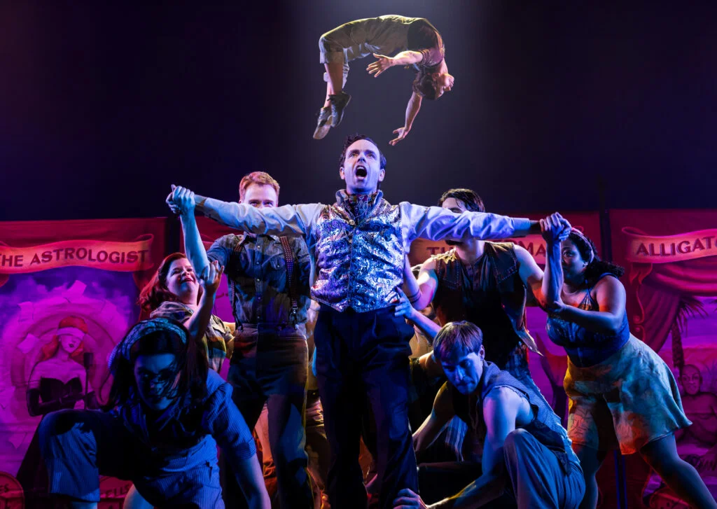 August the ringmaster center stage in his silver vest, arms spread, an acrobat mid-backflip in the air above the company